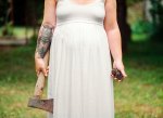 a bigger woman in a white wedding dress holds an axe she has an owl tattoo on her arm she stands outside on the grass