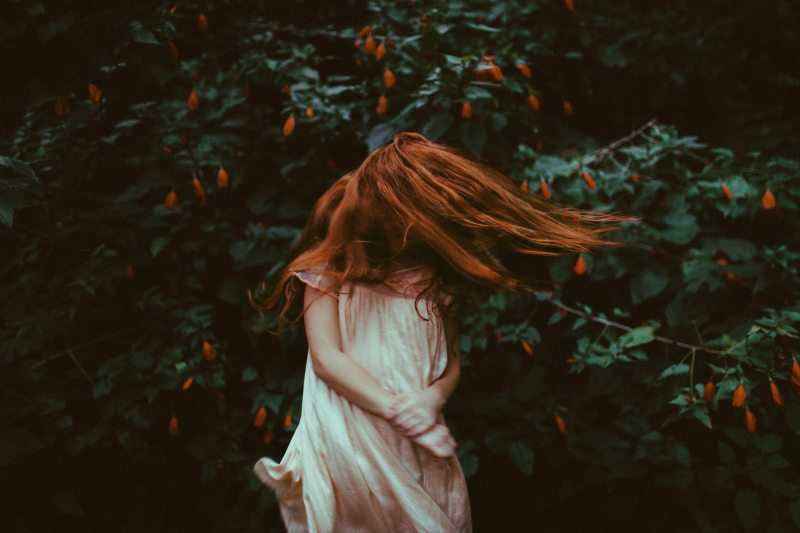 girl with red orange hair wearing a white nightdress swings her hair in front of face while standing in front of a green bush with orange flowers true sex stories tales of sexuality sex confessions